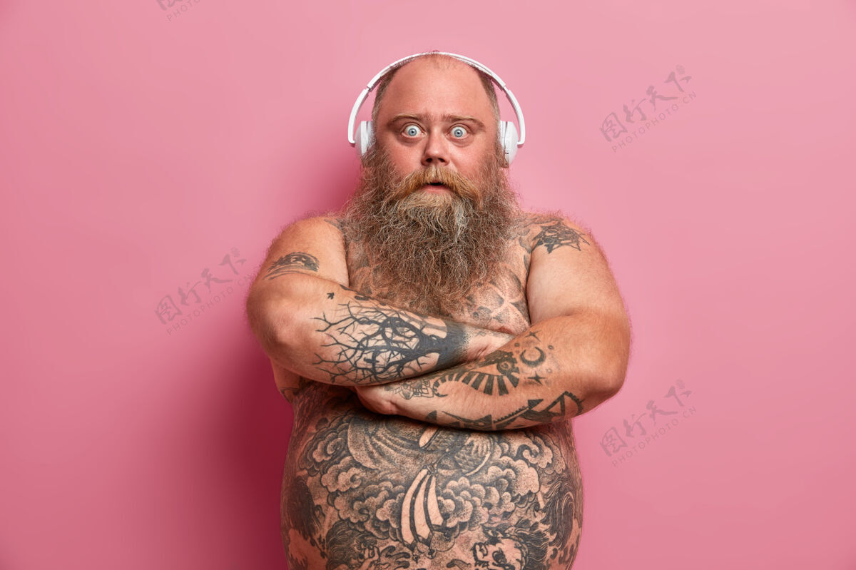 腹部奇怪的胡须男人的肖像保持双臂合拢 用窃听的眼睛看 买了耳机在假日销售 捕捉积极的氛围 有裸体纹身肚子 肥胖 因为懒惰的生活方式男人情绪音乐