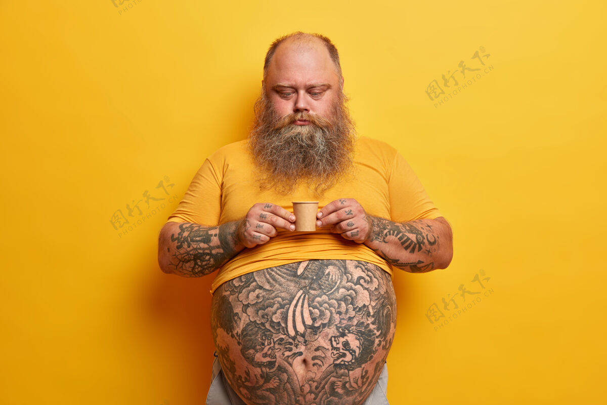 热大肚子 手臂和腹部都有纹身的严肃的大胡子男人 拿着一小盒含糖的咖啡 喝着咖啡因芳香的饮料 穿着黄色t恤 独自在室内摆姿势男人饮用胡子
