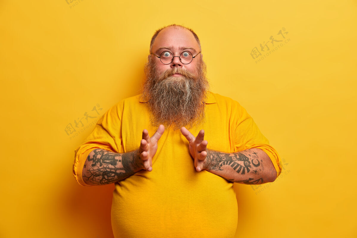 害怕腰部向上拍摄的惊喜胖子有大肚子 眼睛瞪着 举手 害怕什么 穿着休闲t恤 隔离在黄色的墙上超重的惊喜家伙身体胃情绪