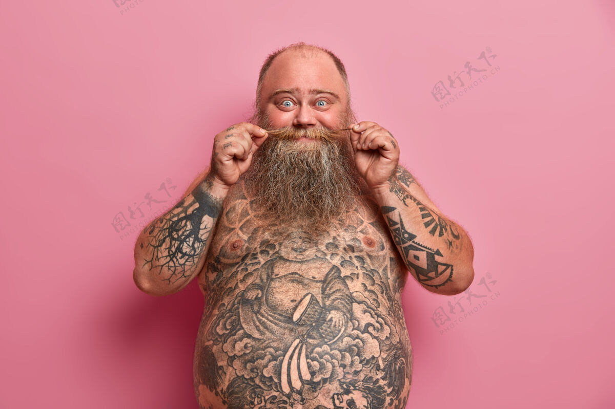 室内搞笑的胡须男人摸胡子 大肚子赤裸着站着 纹身 和朋友聊天 对着粉色的墙摆姿势赤膊上阵的肥胖男人人 营养 体型大站立脂肪