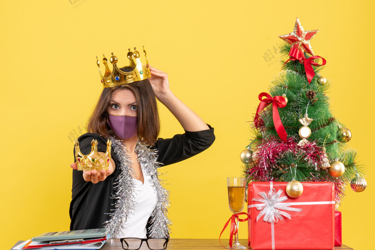 服装圣诞节心情与美丽的女士穿着西装 戴着医用口罩 头上戴着皇冠 手上拿着黄色的办公室头快乐时尚