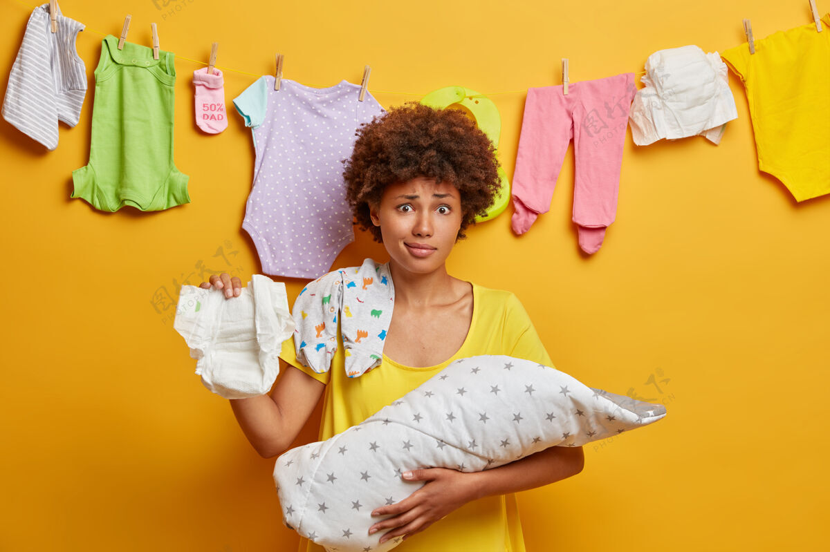 婴儿照顾新生儿困惑的卷发母亲拿着尿布 婴儿在毯子里 忙着护理 给孩子洗衣服 做妈妈的家务 孤立在黄色的墙上妈妈在家里多任务多任务产妇非洲