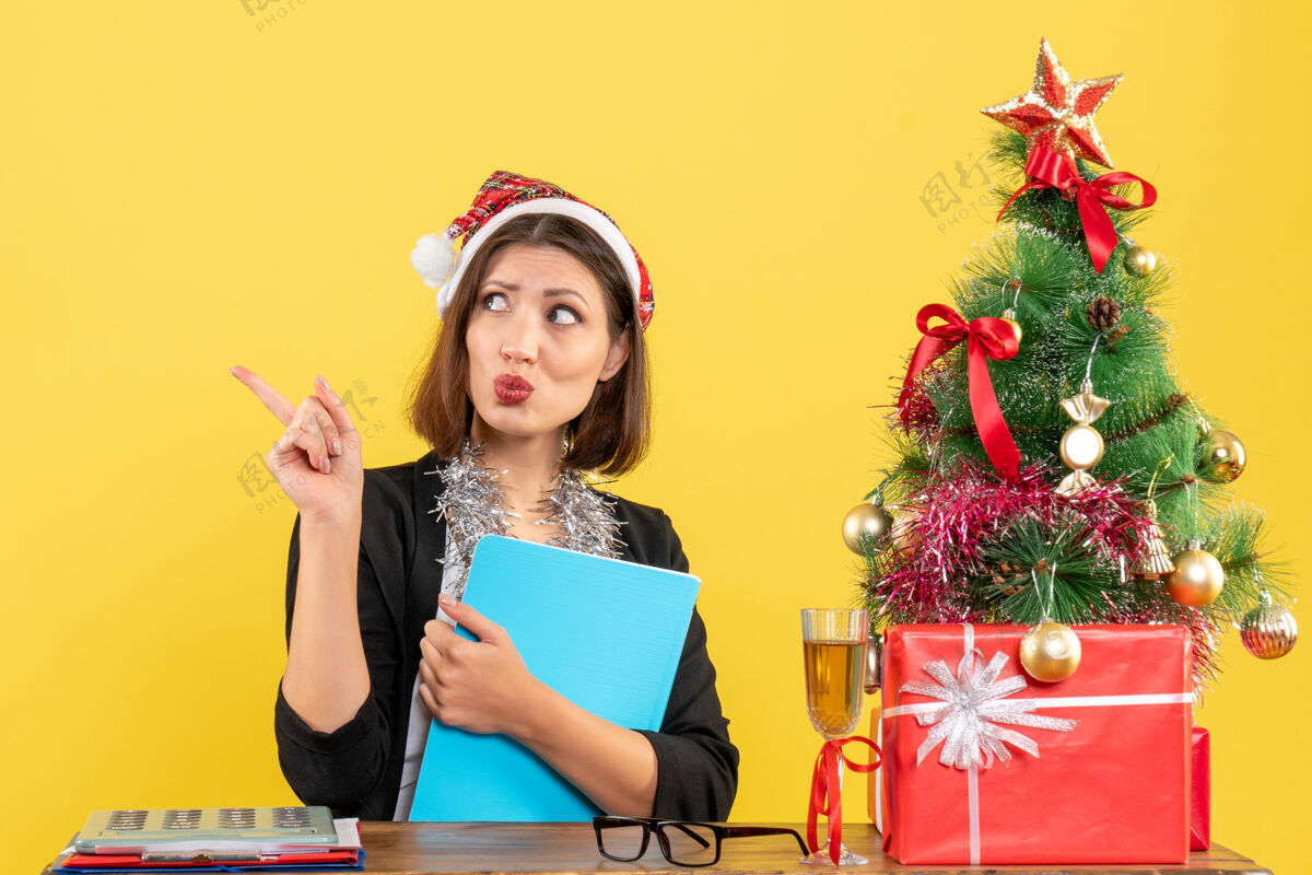 搞笑风趣迷人的女士穿着西装 戴着圣诞老人的帽子和新年的装饰品 手里拿着文件 在办公室里沉思着黄色的孤寂深深时尚人物