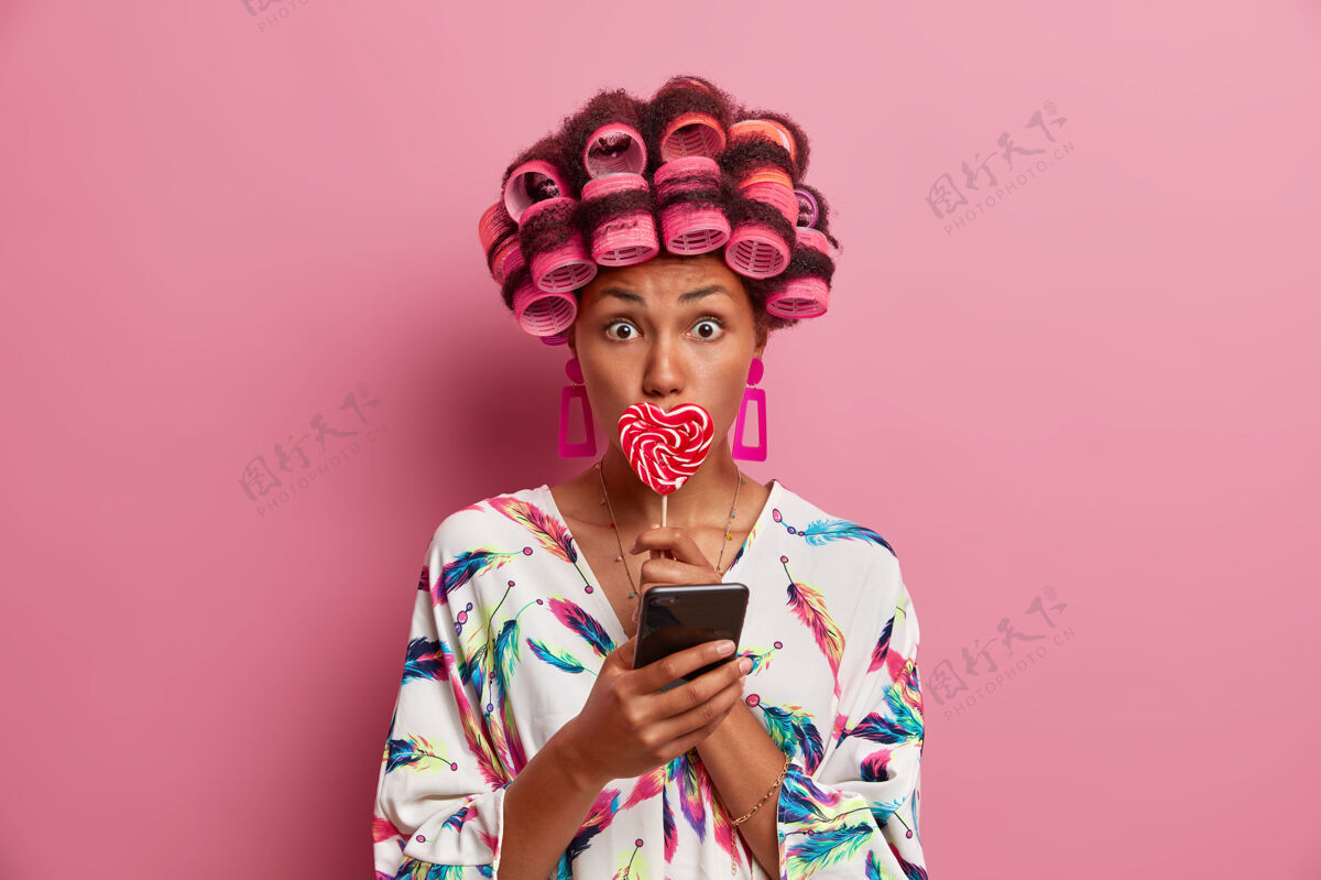 理发师惊讶的年轻女子用卷发器创造美丽的发型 用棒棒糖遮住嘴巴 用智能手机进行在线交流 穿着丝绸睡衣 对着粉色的墙壁摆姿势印象手机电话