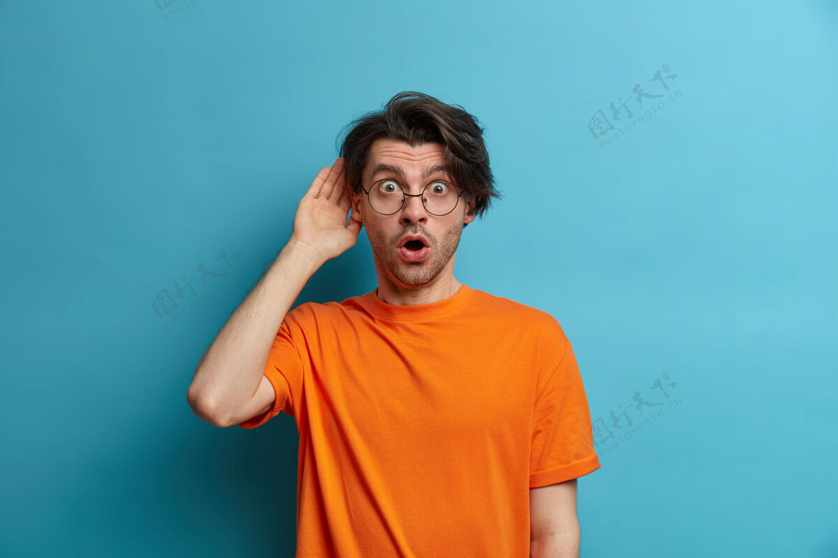 反应人 情感和生活方式的概念惊讶的男人从惊奇中解脱出来 用的眼睛和张开的嘴盯着看 听到可怕的消息 穿着橙色的t恤 戴着透明的眼镜白种人震惊成人