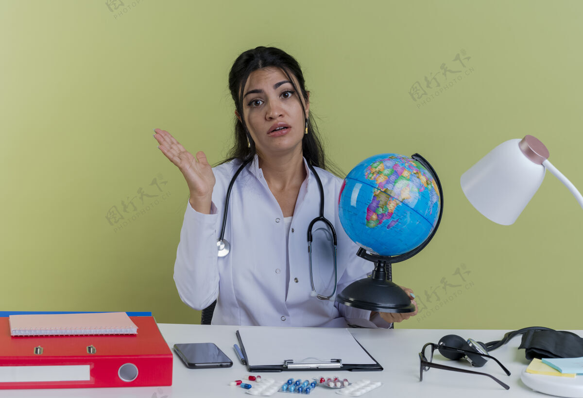 人不知所措的年轻女医生穿着医用长袍 听诊器坐在办公桌旁 手持医疗工具 拿着地球仪 看上去空手孤立桌子医生绿色