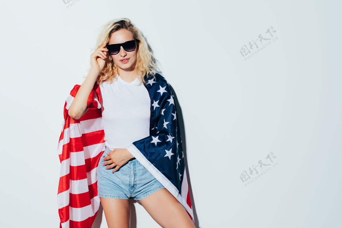 人年轻而无忧无虑年轻漂亮的金发女郎举着美国国旗 微笑着站在白墙上庆祝7月4日独立日模特无忧无虑美国