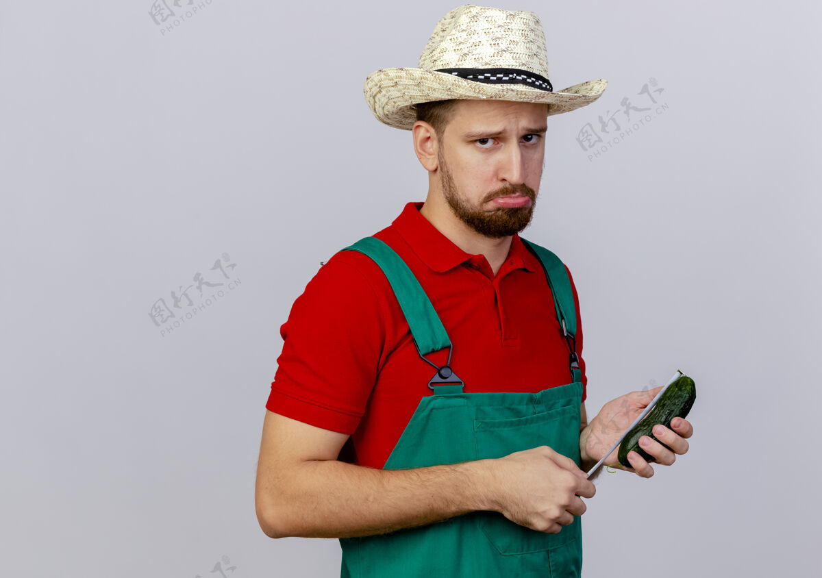 磁带悲伤的年轻英俊的斯拉夫园丁 穿着制服 戴着帽子 手里拿着黄瓜 用卷尺量着隔离的黄瓜年轻测量黄瓜