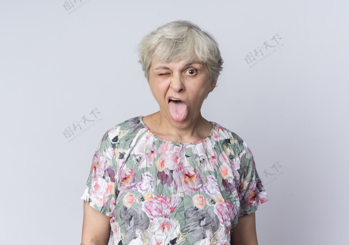 眼睛快乐的老妇人眨着眼睛 伸出舌头孤立地站在白墙上老人女人舌头