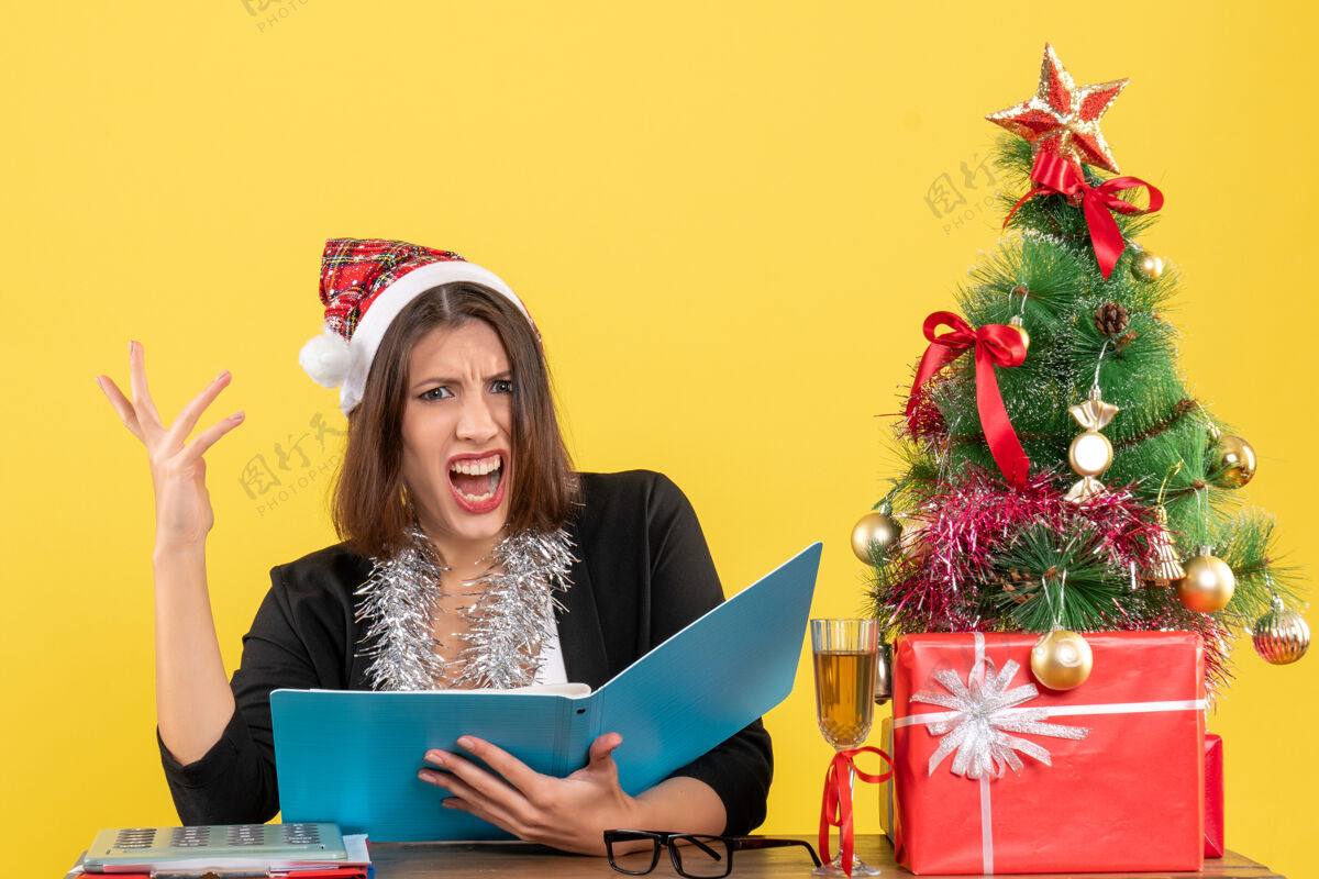 帽子穿着西装 戴着圣诞老人帽和新年装饰品的商务女士专注于文件 感到困惑 坐在办公室的一张桌子旁 桌上放着圣诞树树电脑专注