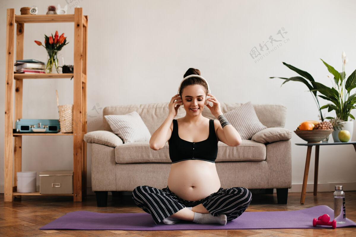 运动在紫色的瑜伽地毯上 快乐的黑发女人戴着耳机听音乐期望怀孕腹部