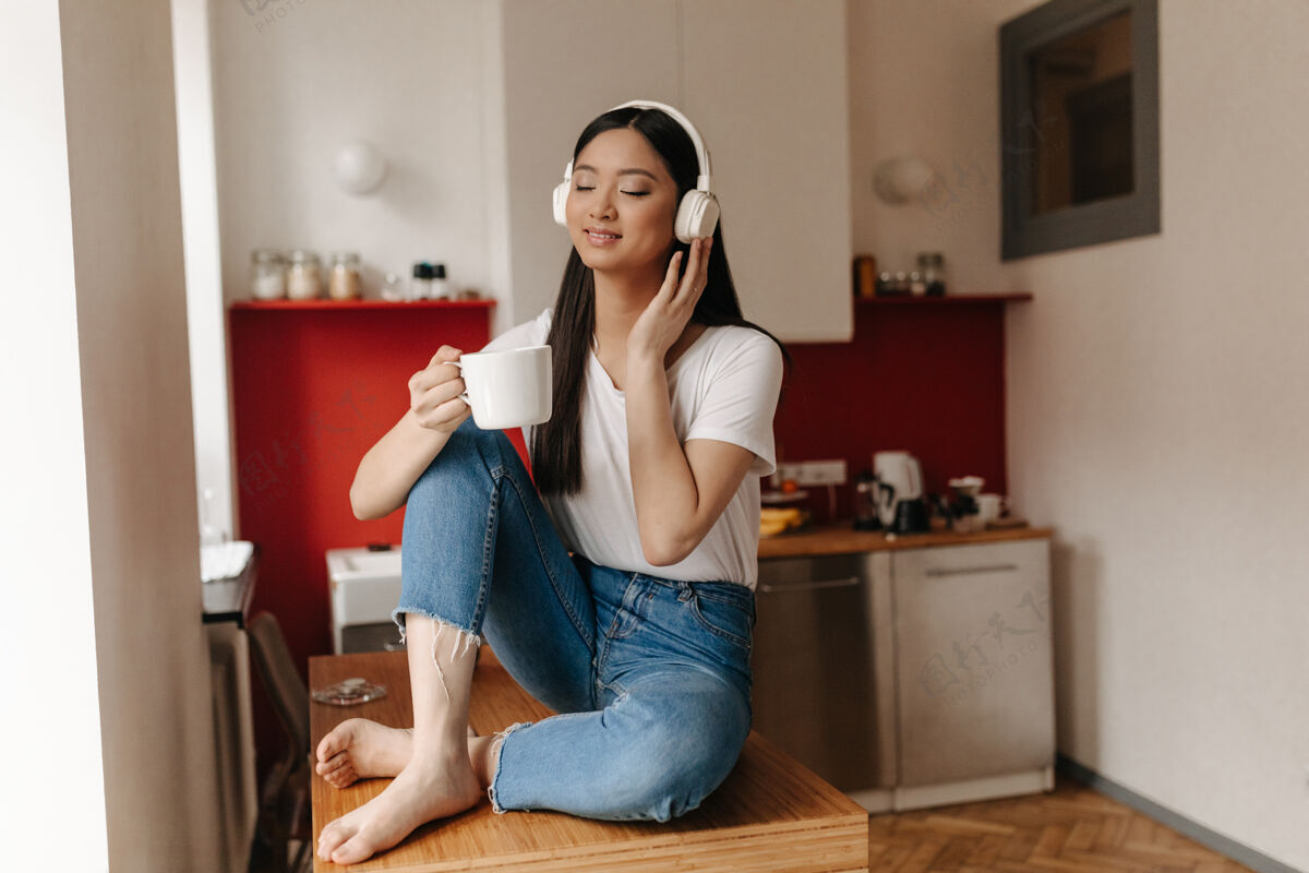 电话穿着牛仔裤和白色上衣的亚洲女人的肖像 戴着耳机在厨房里放松 喝着咖啡女耳机内部