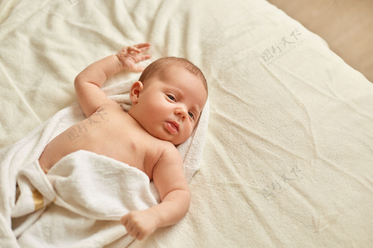 干净刚出生的宝宝沐浴后裹着毛巾躺在床上的白色毛毯上 婴儿望着别处 可爱的孩子洗完澡后皮肤柔软 放松的孩子毛巾洗澡身体
