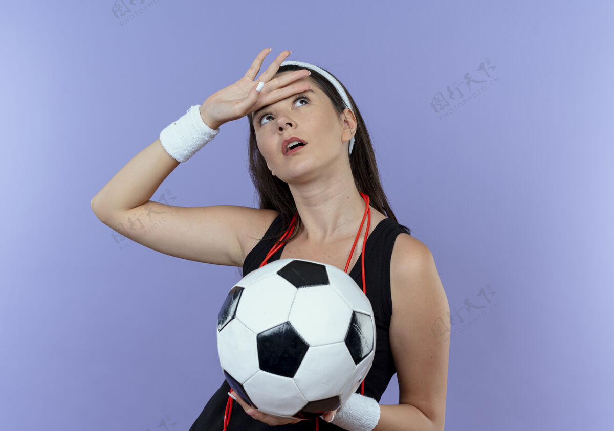 球戴着头带 脖子上系着跳绳 手里拿着足球 站在蓝色背景上看累了的年轻健身女士抱着周围绳子
