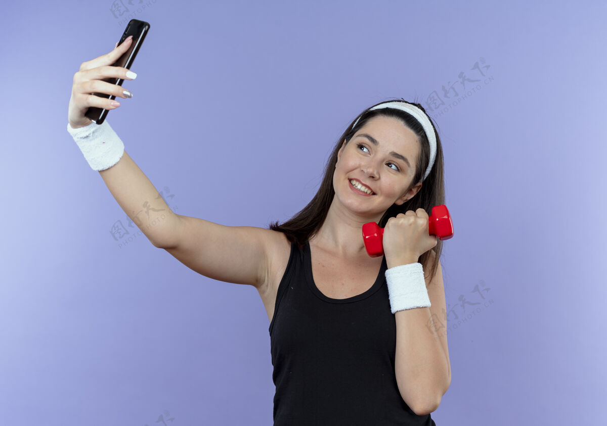 面部戴着头巾的年轻健身女用智能手机自拍手持哑铃微笑着快乐的脸站在蓝色背景上健身微笑抱着