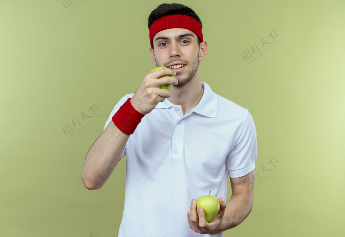 运动戴着头巾的年轻运动型男子手持绿色苹果 站在绿色背景上咬着一个苹果年轻站着