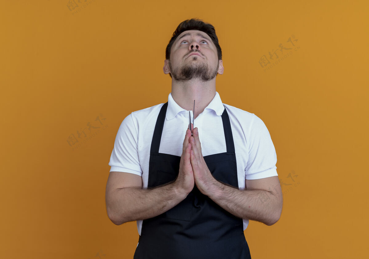 站着围裙里的理发师双手合十 像是在祈祷 站在橙色的背景下 他充满了希望的表情掌心理发师喜欢