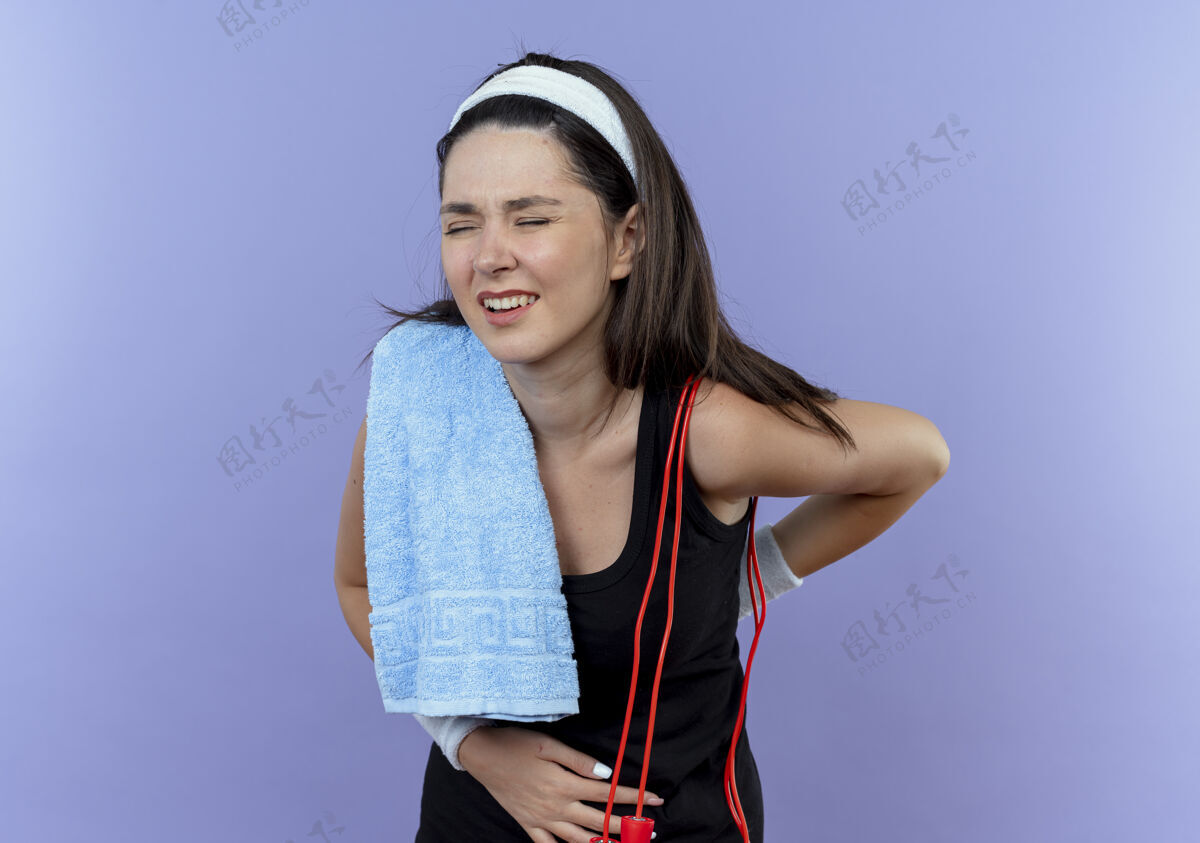 头带戴着头巾 肩上扛着毛巾的年轻健身女士 看起来不舒服 摸着背 站在蓝色背景上感觉疼痛年轻背部站立