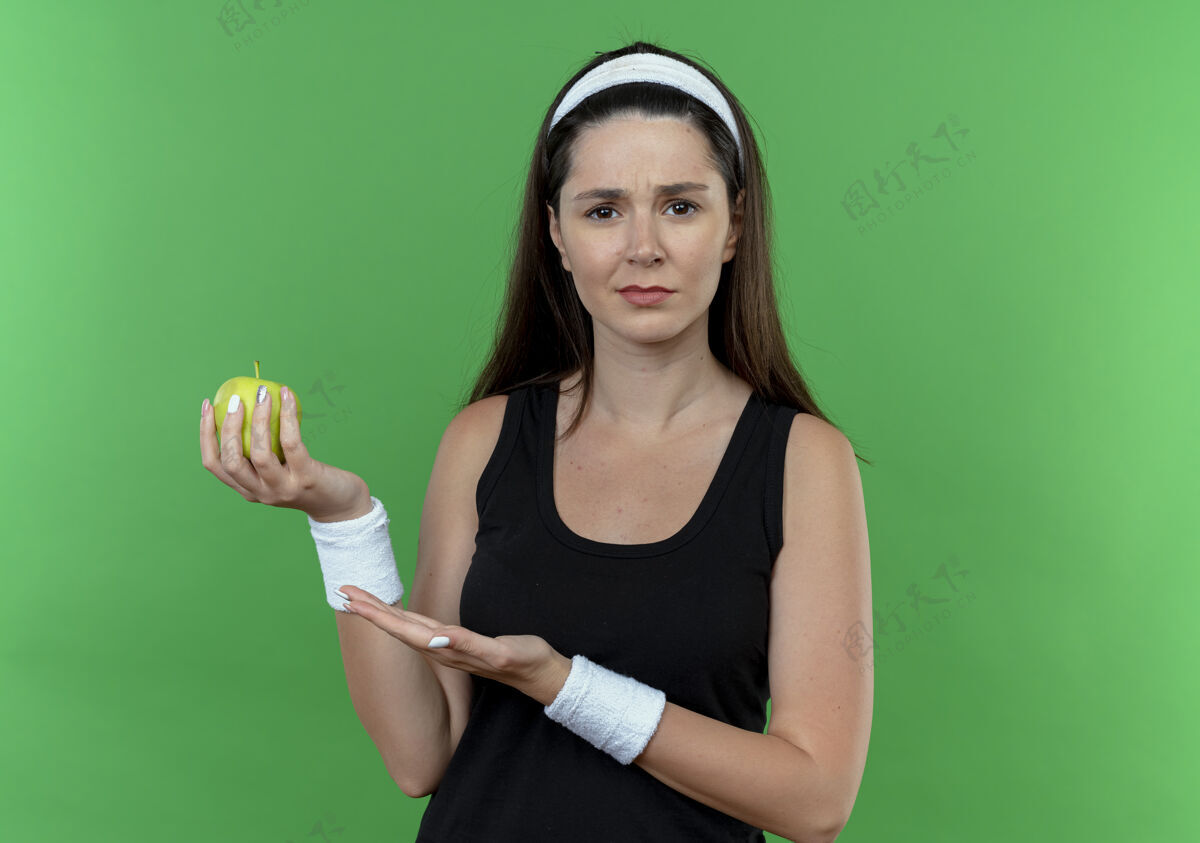 相机戴着头巾的年轻健身女士拿着绿色的苹果站在绿色的背景上困惑地看着相机困惑苹果年轻