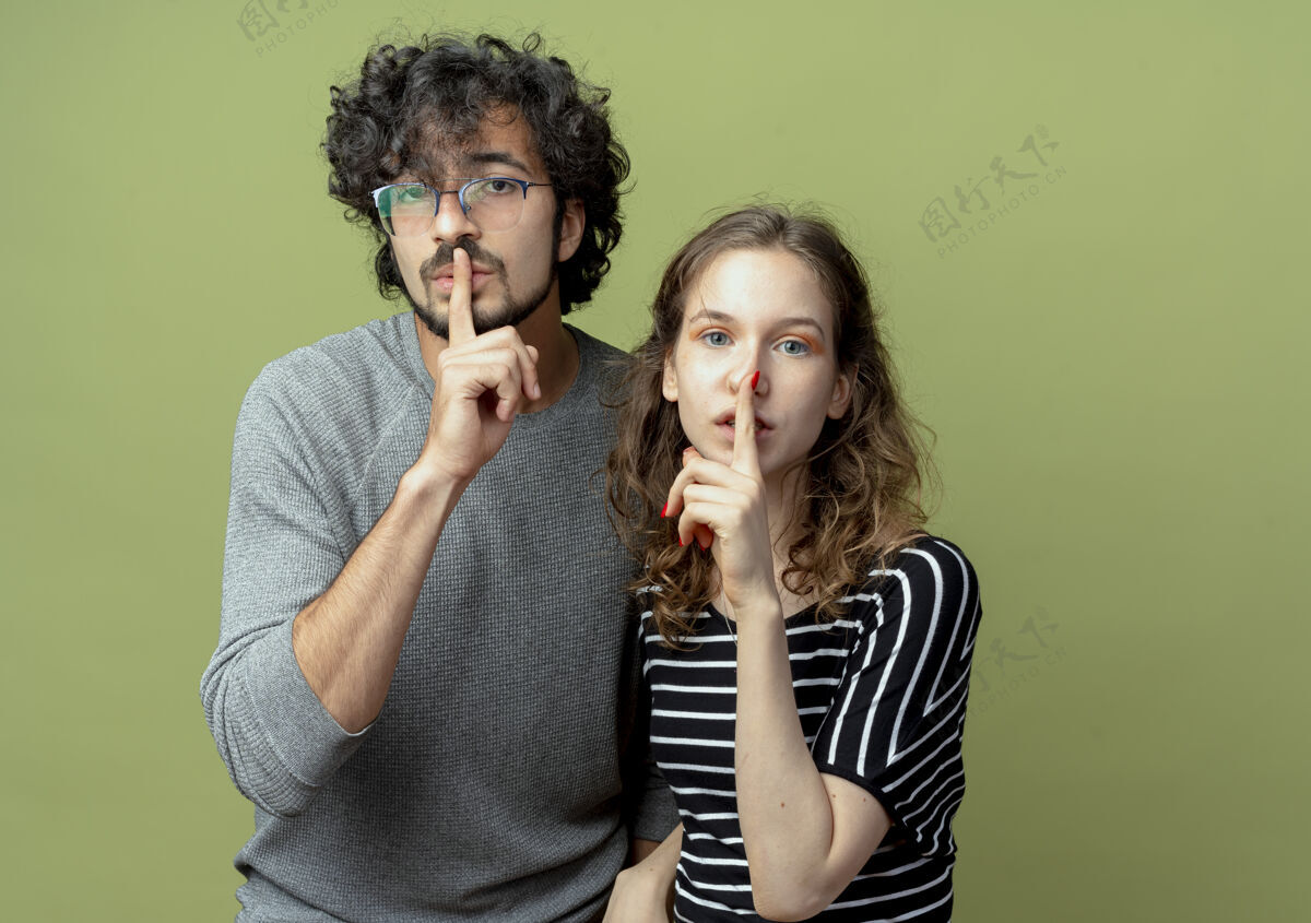 情侣年轻漂亮的情侣们看着相机 在淡绿色的背景下 手指放在嘴唇上做着沉默的手势手势制造年轻