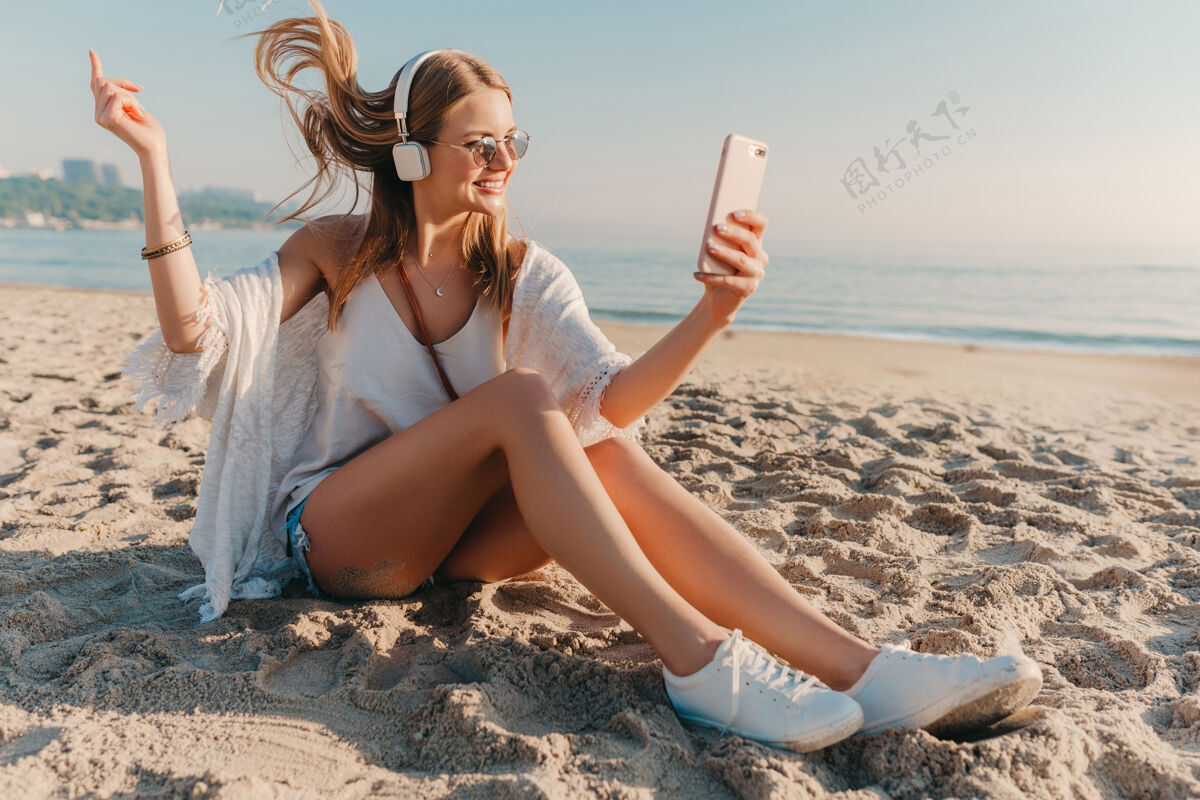 阳光年轻迷人的金发微笑女子在度假时坐在沙滩上打电话自拍沙滩音乐旅程