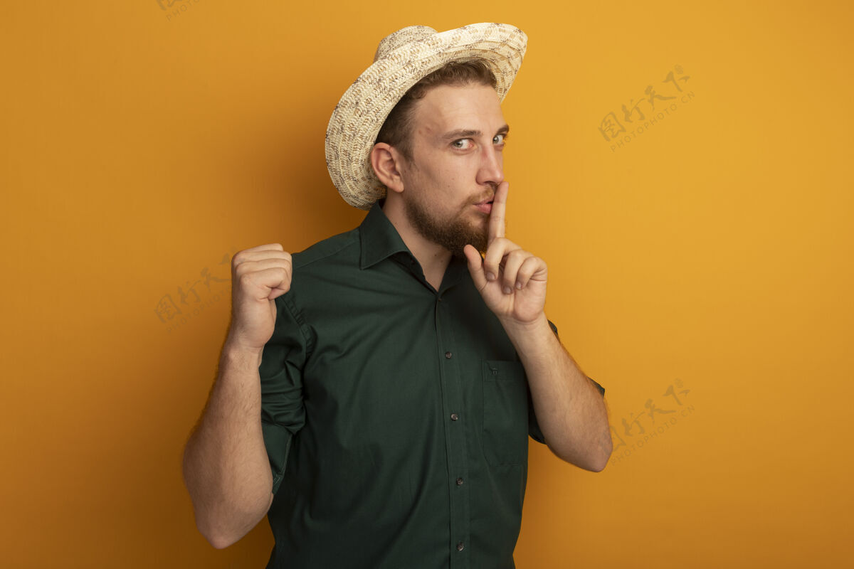 请一个戴着沙滩帽的金发帅哥做着沉默的手势 把拳头孤立在橙色的墙上帽子市民站立