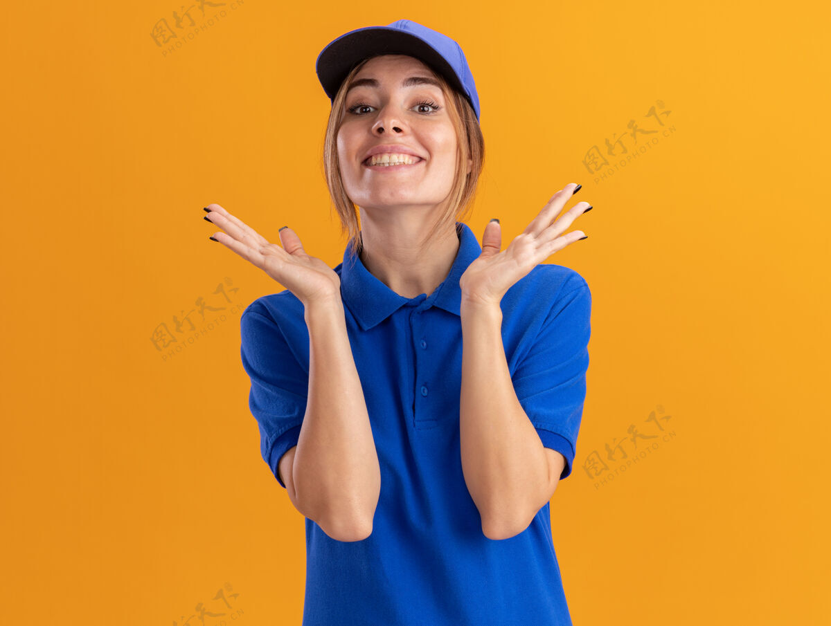 脸微笑着的年轻漂亮的女送货员穿着制服 在橙色的墙上张开双手微笑衣服姿势