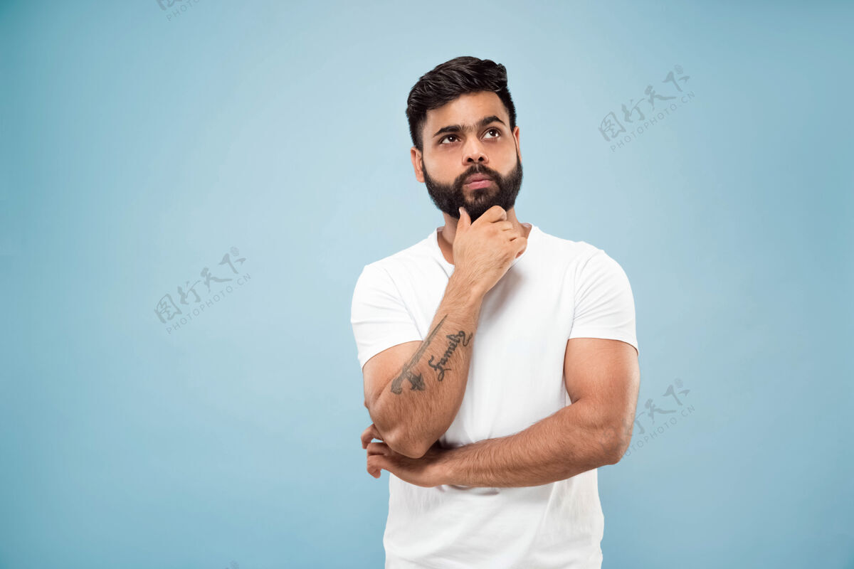 男性蓝色背景上穿着白衬衫的印度教年轻人的半身特写肖像人类情感 面部表情 广告概念负面空间手拉胡子思考选择工作顺序表情