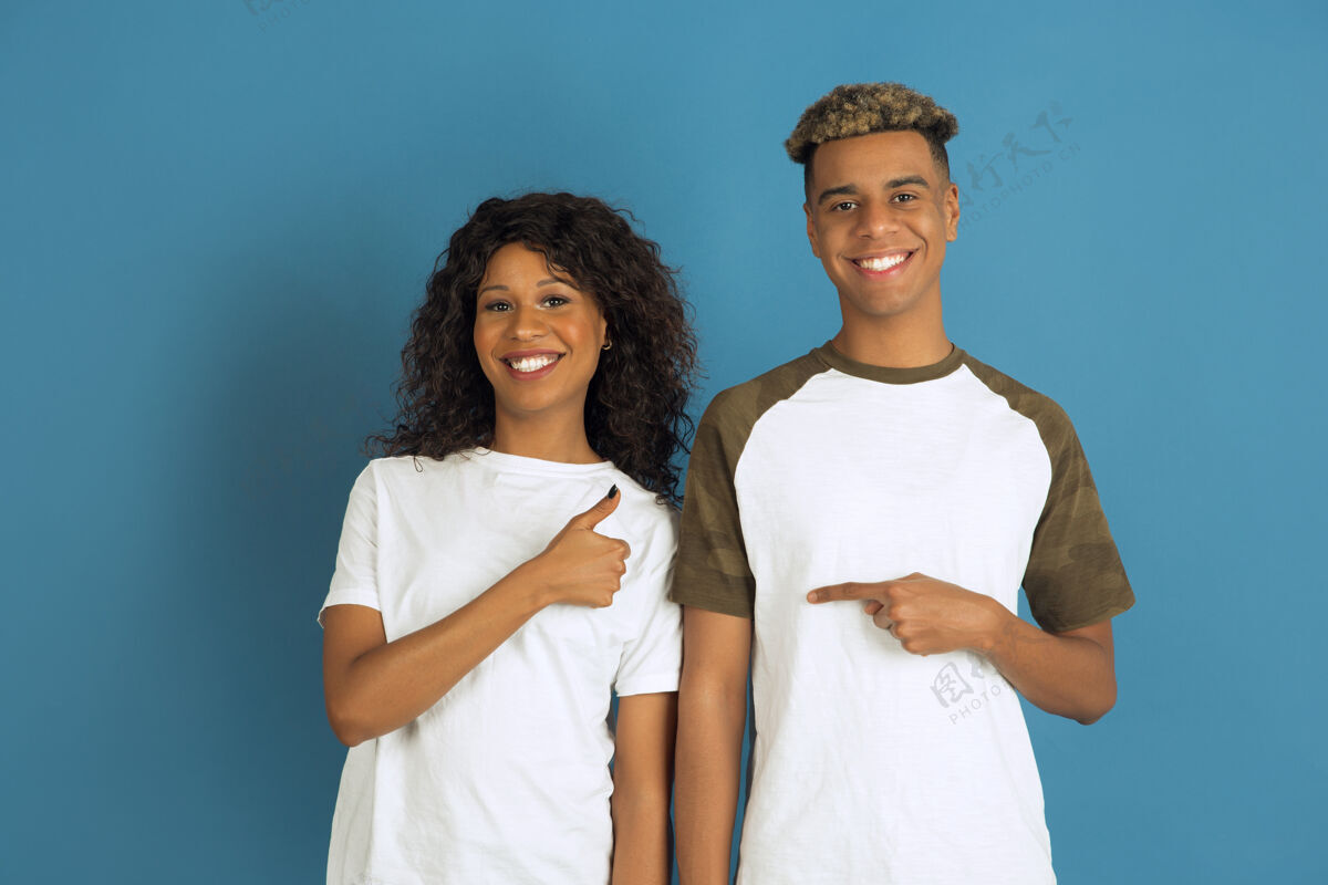 微笑年轻感性的非裔美国人 穿着白色休闲服 在蓝色背景上摆姿势美丽的情侣人类情感的概念 面部表情 关系 互相指指点点的广告姿势笑指向