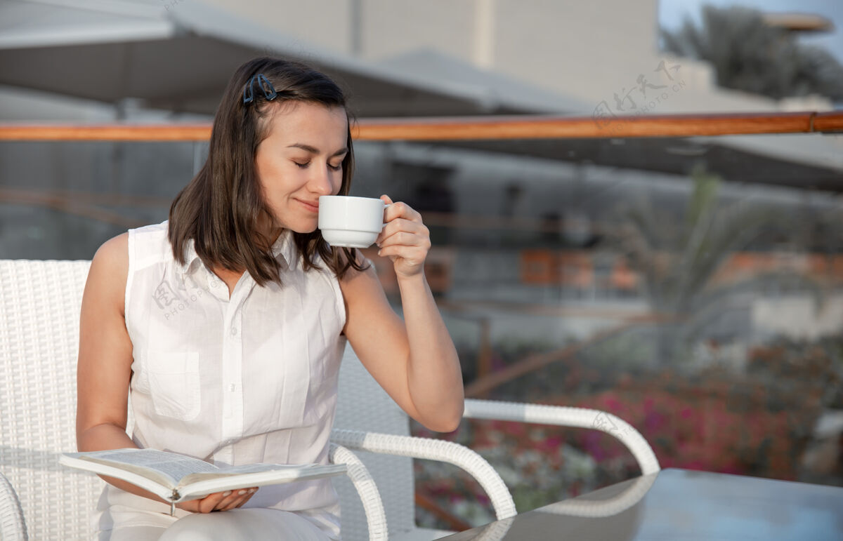 露台年轻的黑发女人手里拿着一杯热饮和一本书享受着早晨的时光休息酒店早晨