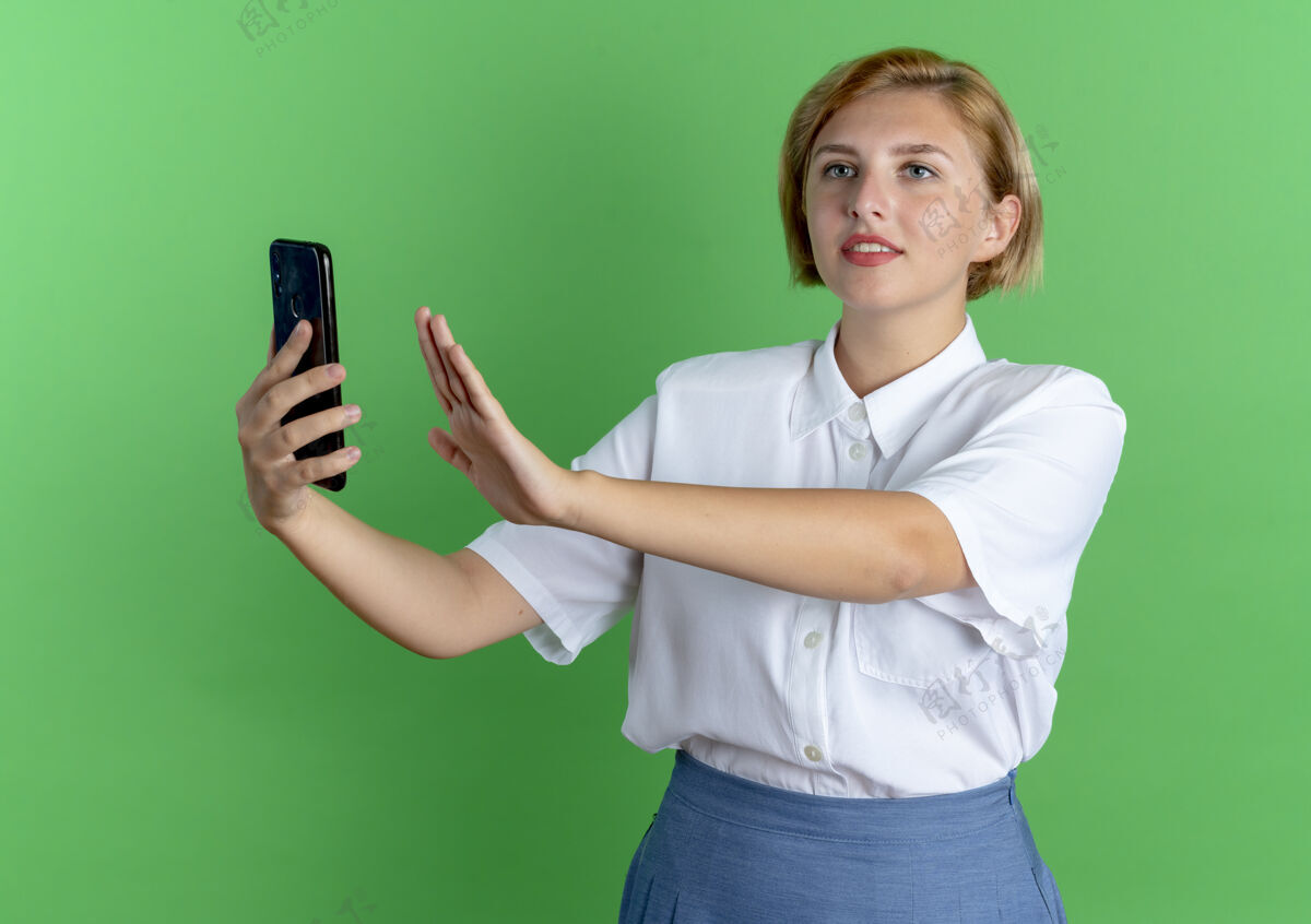 举起来年轻高兴的金发俄罗斯女孩举起手拿着电话 看着绿色背景上有空白的一面俄国人侧线女孩