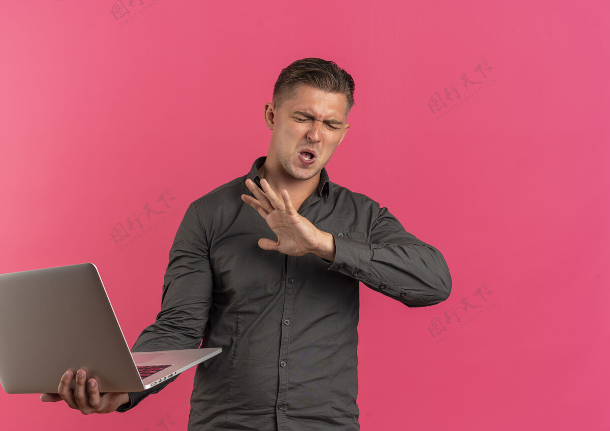 英俊年轻恼怒的金发帅哥拿着笔记本电脑 闭着眼睛 在粉色背景上保持着孤立的手 还有复印空间持有手眼睛