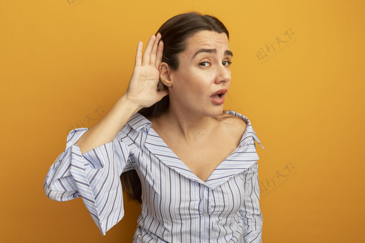 姿势恼怒的漂亮女人把手放在耳朵后面 试图听到橘色墙上的孤立声音烦恼感觉站着