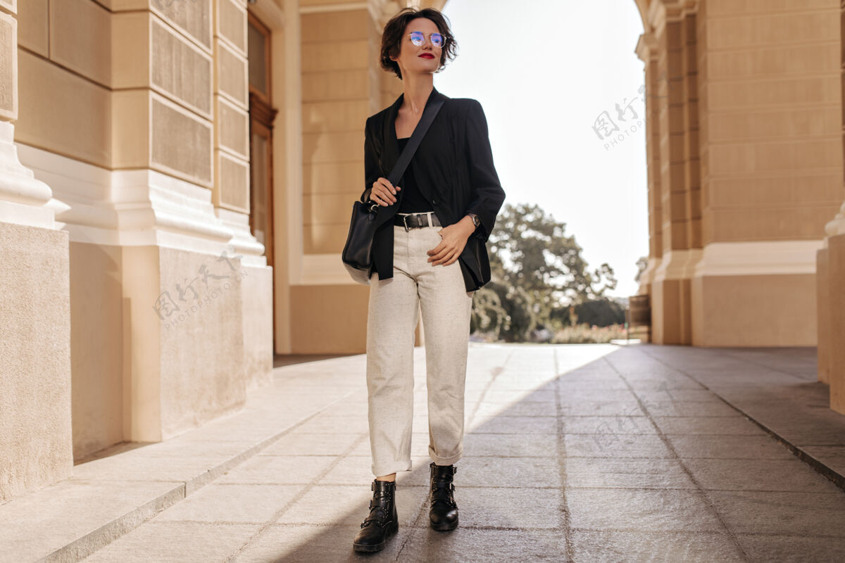 模型穿着轻便长裤 靴子和夹克的女人在外面摆姿势的全身照片戴着黑色手提包和眼镜的迷人女人在街上微笑眼镜姿势步行
