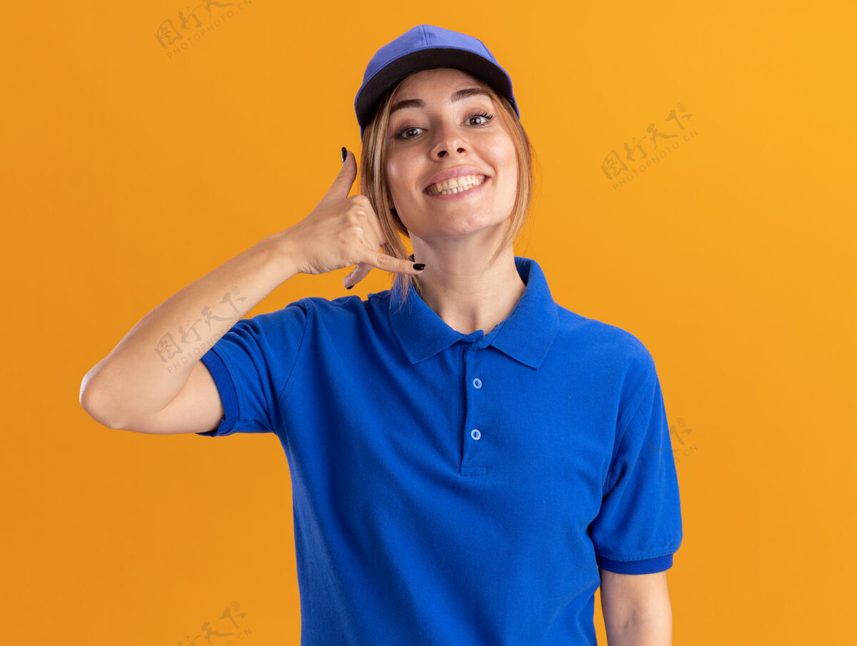 年轻微笑着的年轻漂亮的女送货员 穿着制服 手势叫我 手势孤立在橙色的墙上人姿势电话