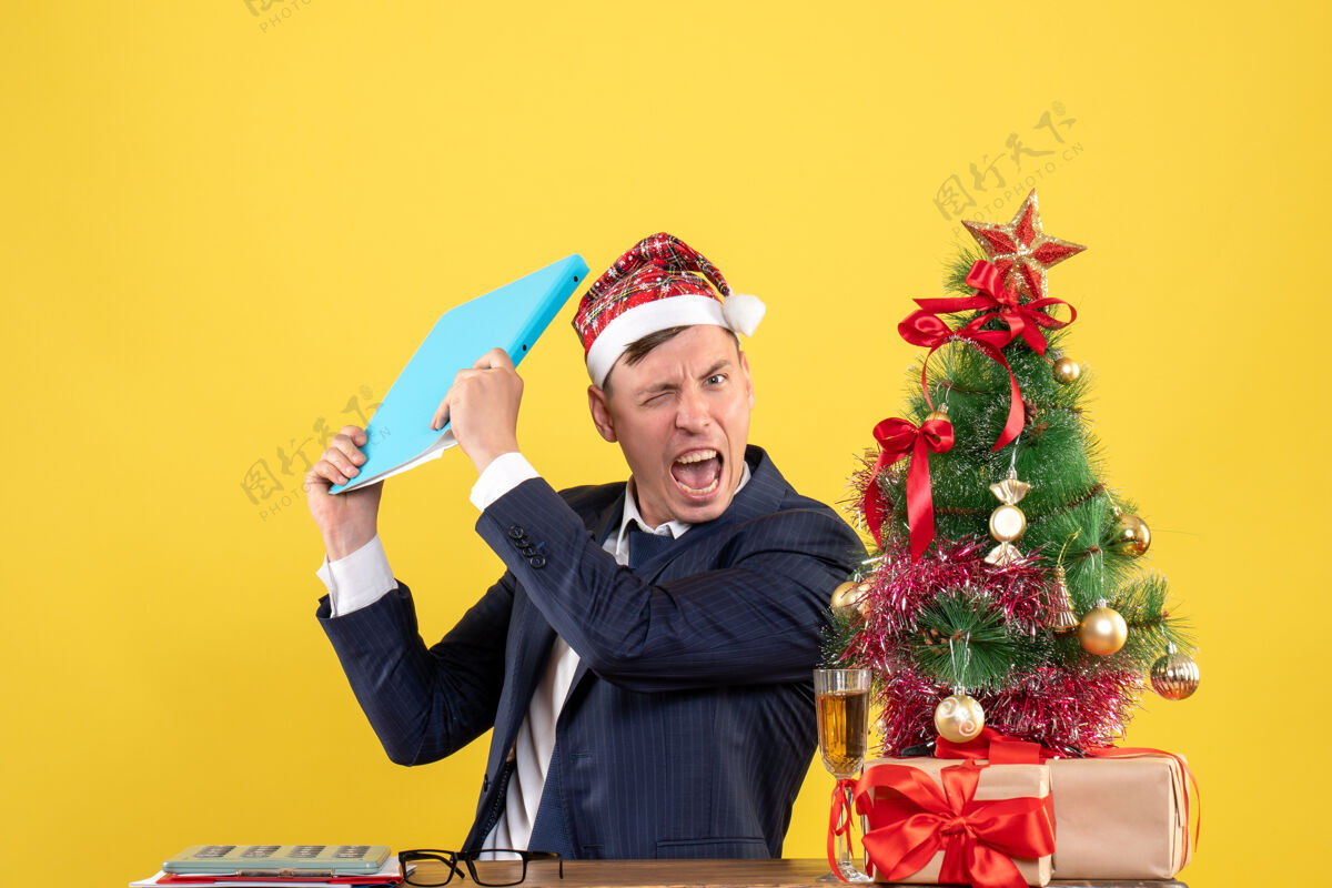 男人前视图愤怒的商人拿着文件文件坐在圣诞树旁的桌子旁 黄色背景下呈现人坐着人