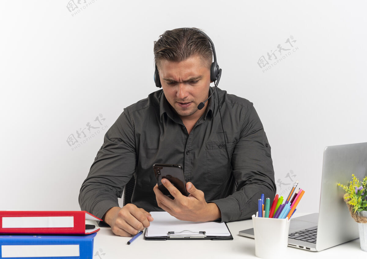 使用年轻恼怒的金发上班族戴着耳机坐在办公桌旁 拿着办公工具用笔记本电脑看着白色背景上的手机 还有复印空间金发工具工作人员