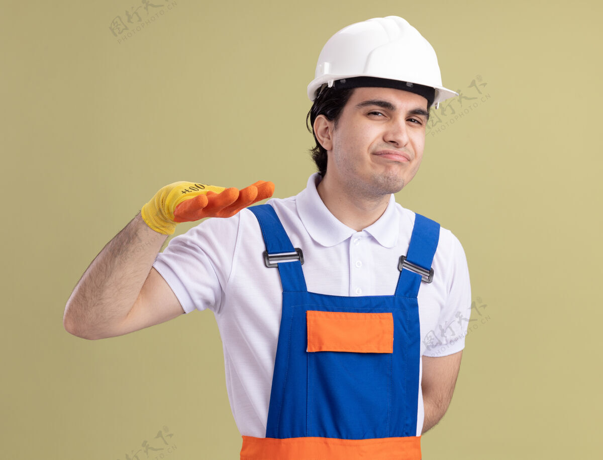 建筑工人年轻的建筑工人穿着建筑制服 戴着安全帽 戴着橡胶手套 站在绿色的墙上 看着前面做着冷静的手势放下站立建筑
