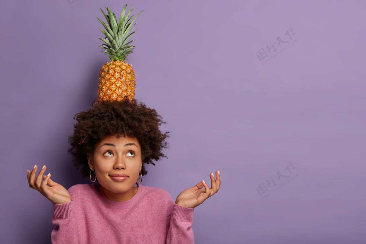 热带黑色卷发女人的水平视图试图保持平衡 将新鲜菠萝放在头上 摊开手掌 向上看空白犹豫菠萝