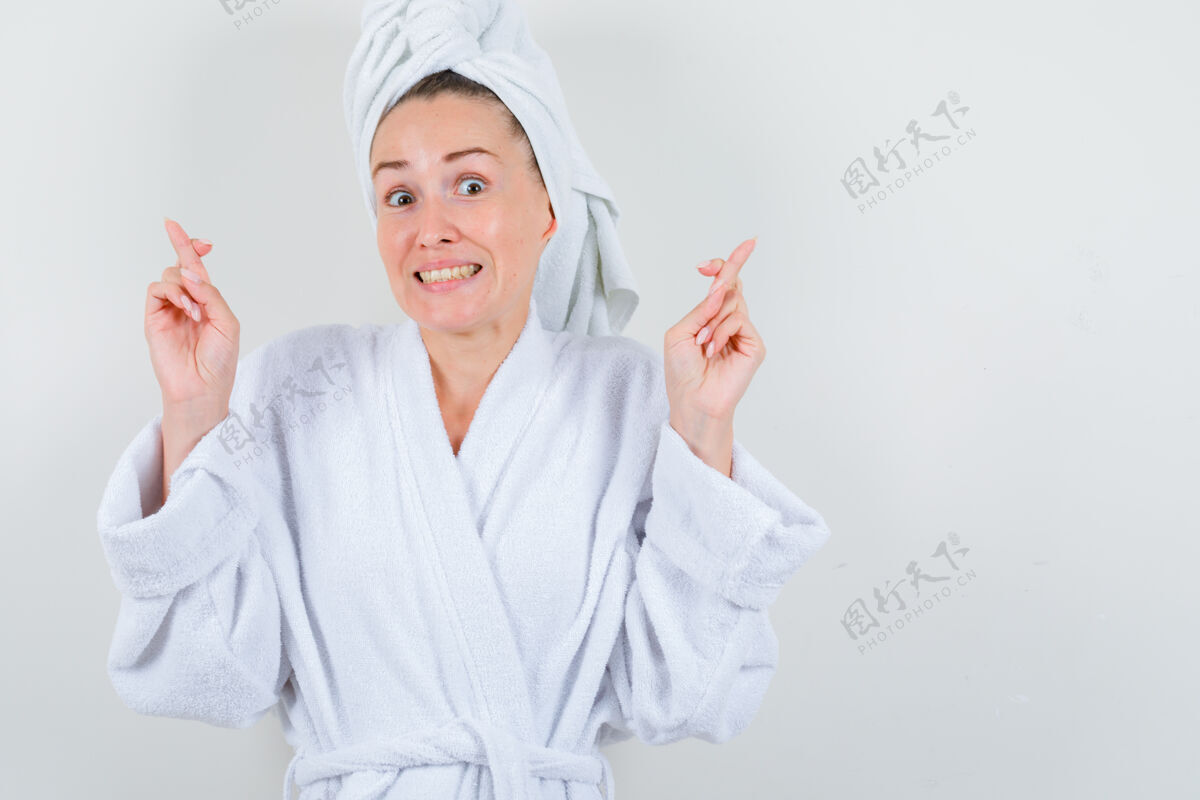 人年轻的女士在白色浴衣 毛巾和保持手指交叉 看起来很高兴 前视图护理皮肤保持