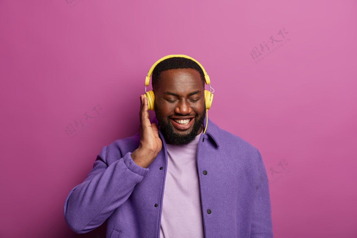 美国微笑的黑人戴着耳机享受美妙的声音 制作新的播放列表 在业余时间听喜爱的音乐 穿着紫色夹克 露出洁白的牙齿乐观休闲收音机