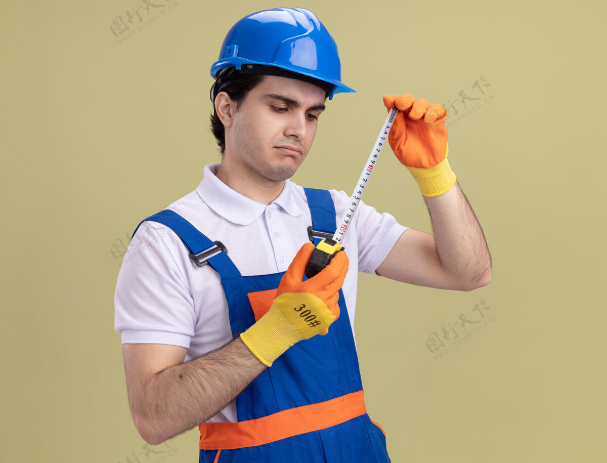 头盔年轻的建筑工人穿着建筑制服 戴着安全帽 戴着橡胶手套 拿着卷尺 严肃地站在绿色的墙上看着它站着脸手套