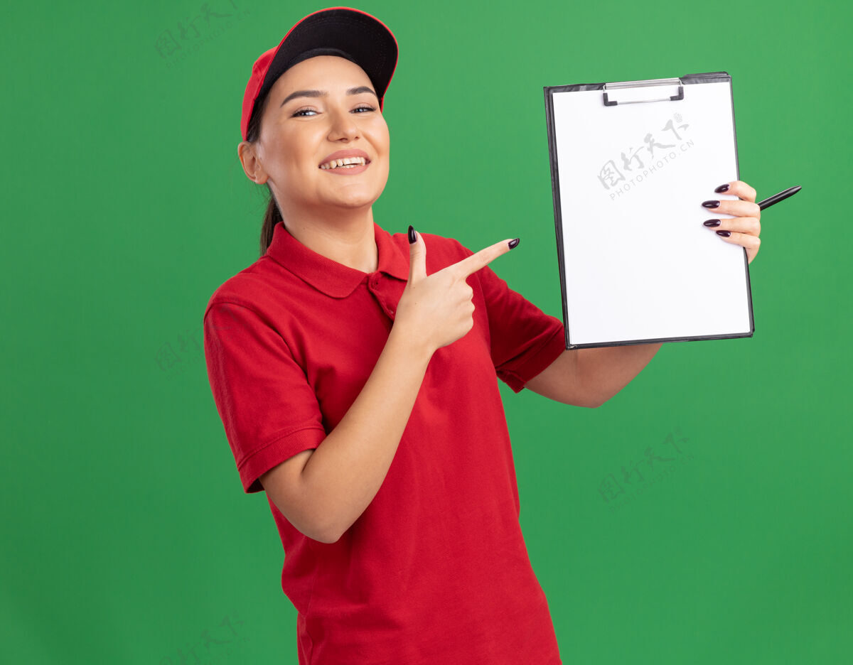 制服身穿红色制服 头戴鸭舌帽的年轻女送货员拿着空白页的剪贴板 用食指指着剪贴板 微笑着看着前面站在绿色的墙上欢呼站着女人