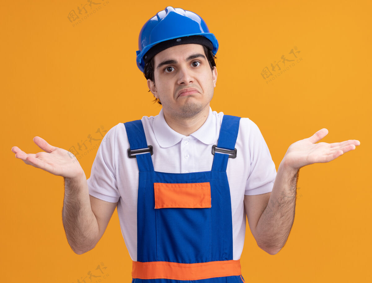 不确定年轻的建筑工人穿着建筑制服 戴着安全帽 站在橙色的墙上 困惑而不确定地耸耸肩看着前方建设者年轻制服