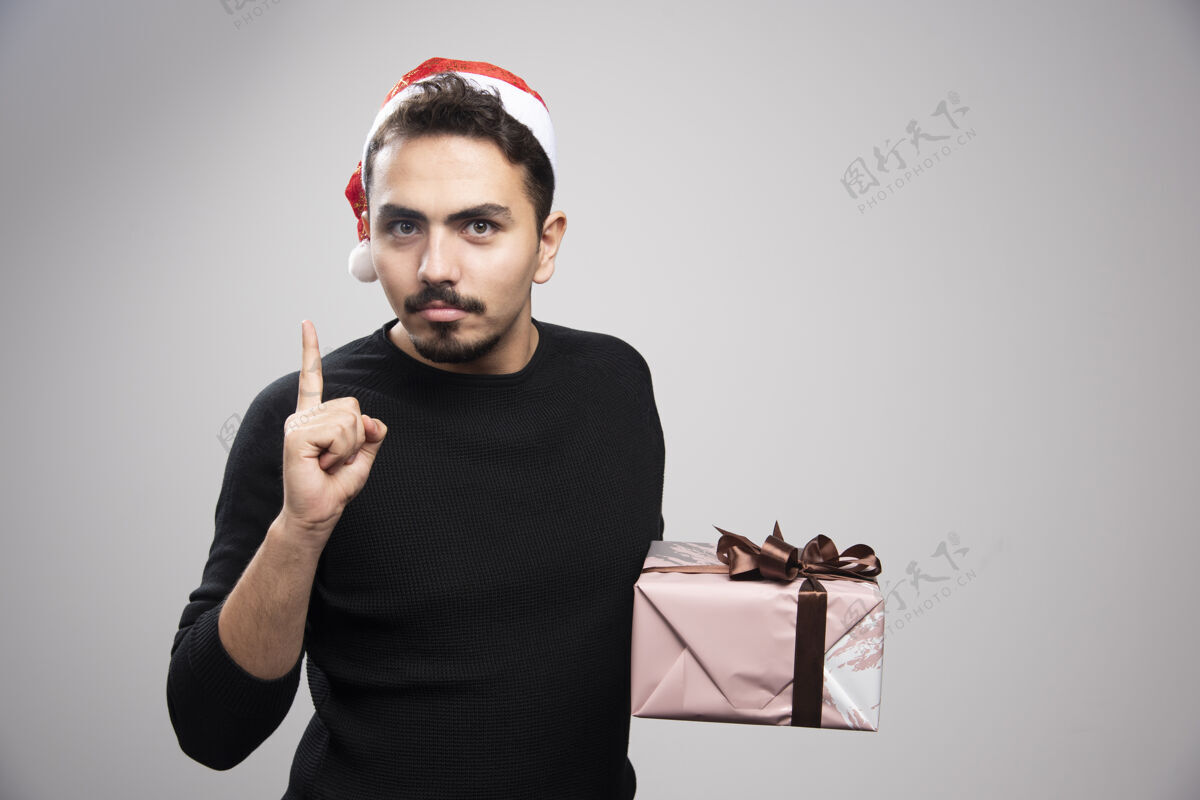 盒子一个戴着圣诞老人帽子的年轻人举起手指拿着礼物毛衣情感模特