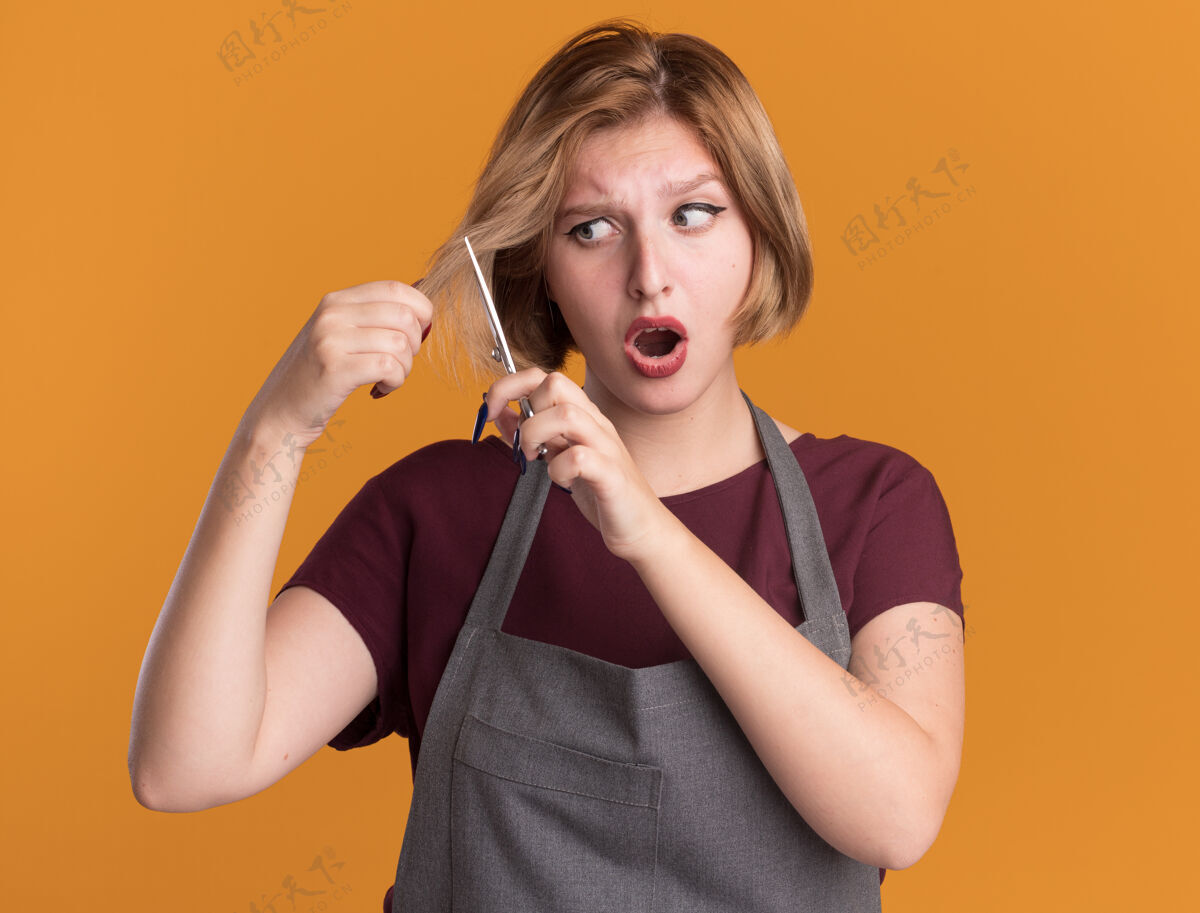 剪年轻漂亮的女理发师站在橘色的墙上 围着围裙试图用剪刀剪头发 看上去既困惑又惊讶理发师剪刀站