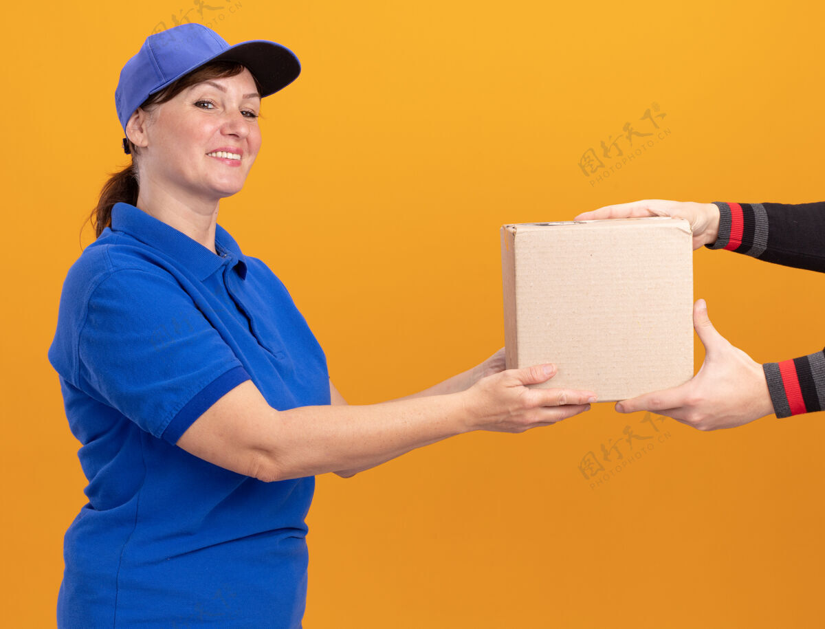 制服身着蓝色制服 头戴帽子的中年送货妇女站在橙色的墙上 微笑着向顾客赠送纸板箱年龄站立赠送