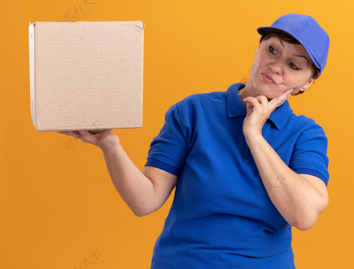 女人中年送货员身穿蓝色制服 戴着帽子 手里拿着纸板箱 站在橘色的墙上困惑地看着它制服拿着中间