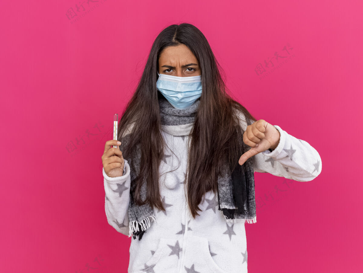 面具戴着医用面罩 戴着围巾 拿着温度计 拇指朝下 粉红色背景下 一个未出狱的年轻女孩年轻显示医疗