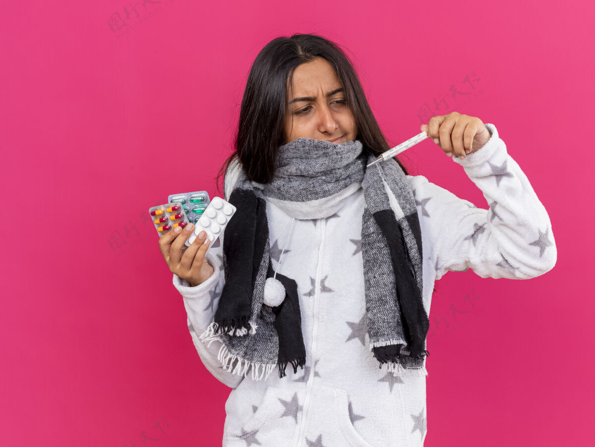 疾病未出狱的年轻病女孩戴着围巾捂着嘴拿着药片 看着手上的温度计孤立在粉红色的背景上体温计女孩药丸